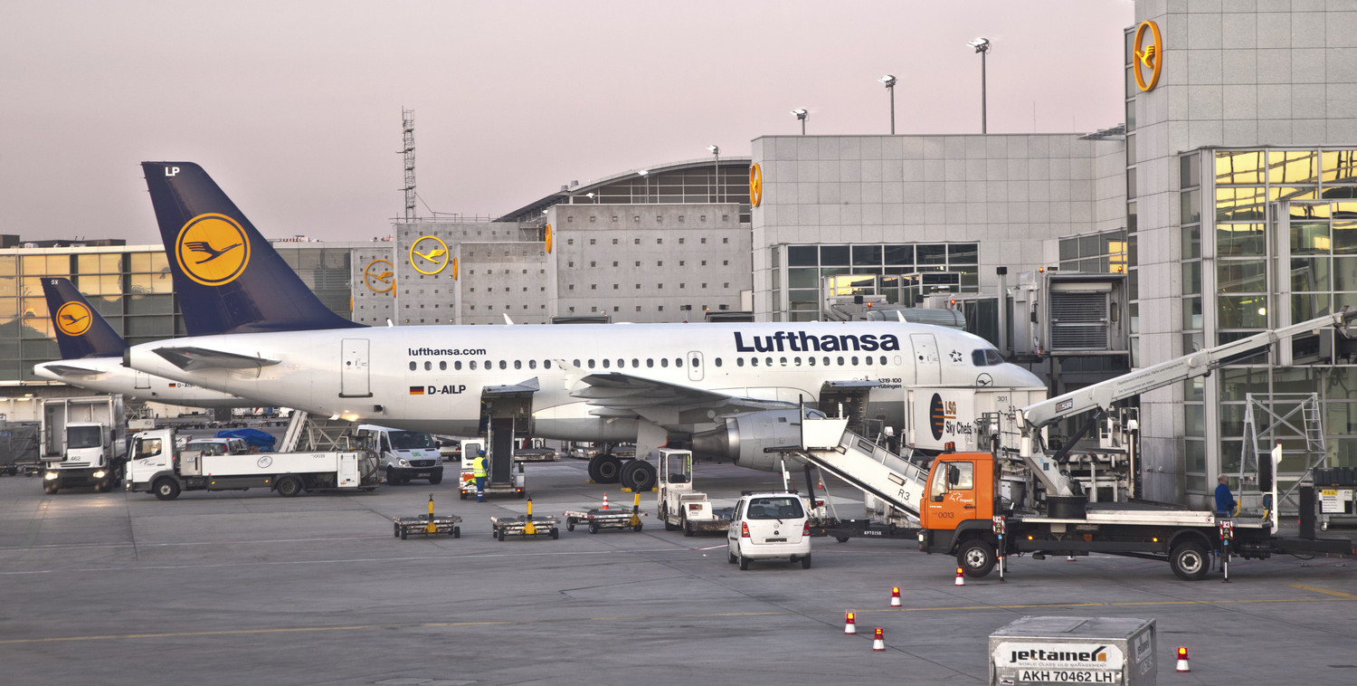 KI generiert: Das Bild zeigt ein Lufthansa-Flugzeug, das am Gate eines Flughafens geparkt ist, während es von verschiedenen Bodenfahrzeugen und -personal betreut wird. Im Hintergrund sind Flughafenarchitektur und weitere Flugzeuge zu sehen.