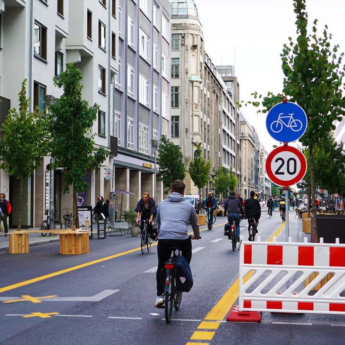 Fahrräder fahren zwischen Häuserzeilen auf einer Pop-Up Fahrradstraße. Sie befindet sich auf einer breiten Straße und wird von gelben Linien begrenzt. 