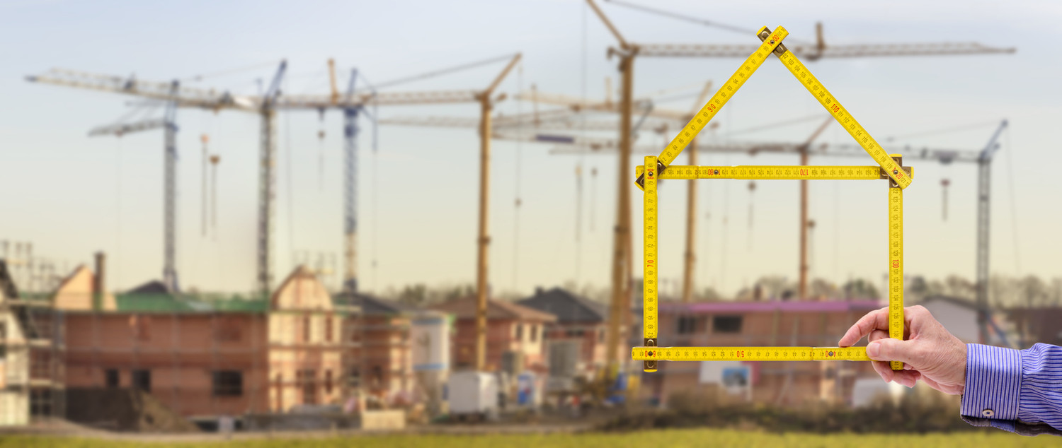 KI generiert: Das Bild zeigt eine Hand, die einen Zollstock in Form eines Hauses hält, im Vordergrund, während im Hintergrund Baukräne und im Bau befindliche Gebäude zu sehen sind. Der Hauptinhalt des Bildes ist das Thema Hausbau und Konstruktion.