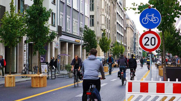 KI generiert: Das Bild zeigt eine belebte Straße, die für Radfahrer freigegeben ist, mit zahlreichen Menschen, die darauf Fahrrad fahren. Verkehrsschilder deuten auf eine Geschwindigkeitsbegrenzung von 20 km/h für Fahrräder hin.