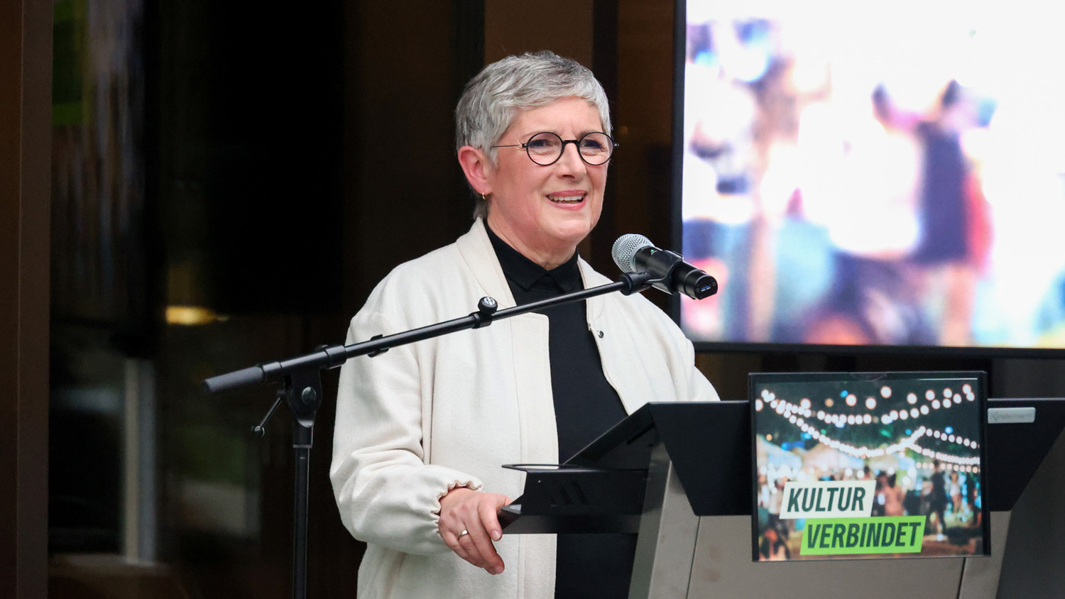KI generiert: Das Bild zeigt eine Frau, die vor einem Mikrofon an einem Rednerpult steht und spricht. Neben ihr befindet sich ein Schild mit der Aufschrift "Kultur verbindet".