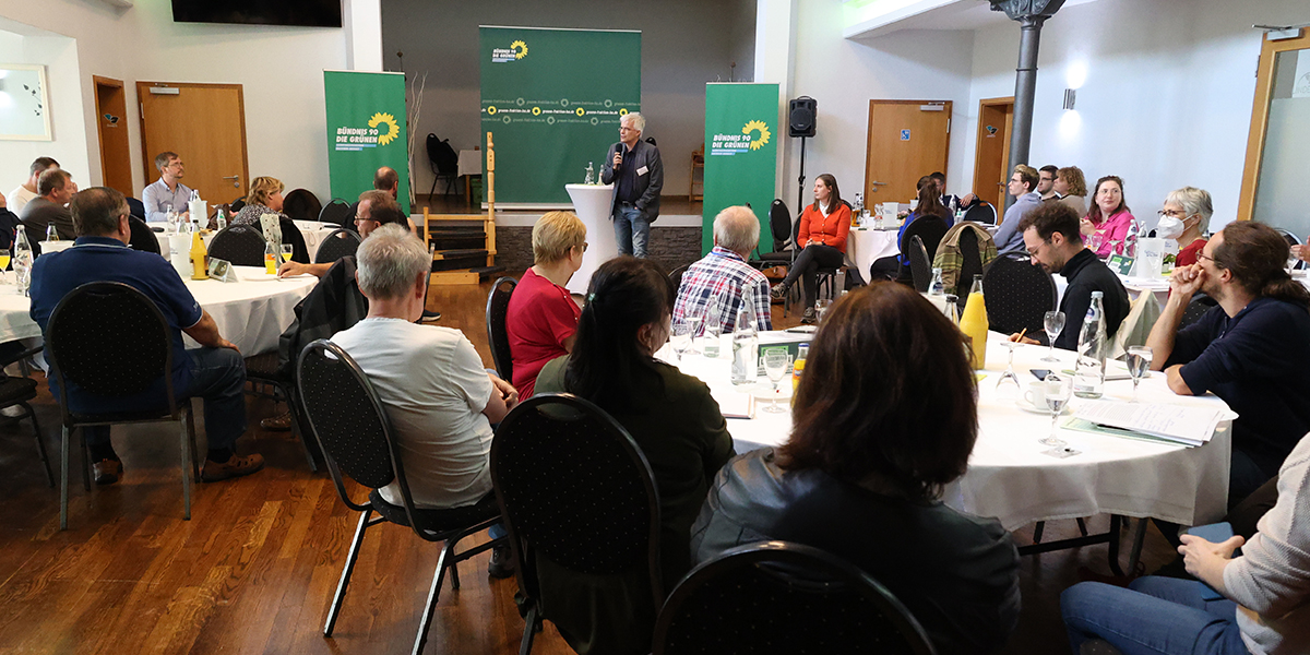 KI generiert: Das Bild zeigt eine Versammlung in einem Konferenzraum, in dem mehrere Leute an runden Tischen sitzen und einem Vortragenden zuhören. Im Hintergrund sind grüne Banner mit einem Logo zu sehen.