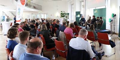 KI generiert: Das Bild zeigt ein Indoor-Event, bei dem eine Gruppe von Personen in einem Publikum sitzt und einer Paneldiskussion oder Präsentation zuhört. Im Vordergrund sind mehrere Menschen in Stuhlreihen platziert, während im Hintergrund Diskussionsteilnehmer auf einer Bühne sitzen.