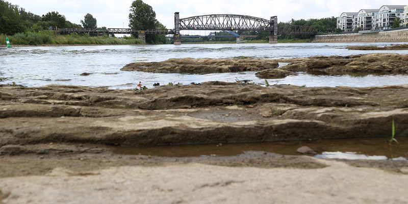 KI generiert: Das Bild zeigt eine flache, felsige Flusslandschaft mit einer Stahlbrücke im Hintergrund, die den Fluss überquert. An den Ufern sind Vegetation und Gebäude sichtbar.