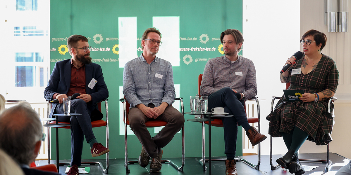 KI generiert: Das Bild zeigt eine Podiumsdiskussion mit vier Personen, die auf einer Bühne sitzen. Im Hintergrund ist ein Banner mit einem grünen Logo und der Aufschrift "gruene-fraktion-lsa.de" zu sehen.