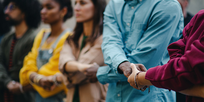 KI generiert: Das Bild zeigt eine Gruppe von Menschen, die sich an den Händen halten und eine Art Kette bilden, was auf Solidarität oder gemeinsames Engagement hinweisen könnte. Sie stehen in einer Reihe, teilweise unklar im Hintergrund, was auf Bewegung oder eine Versammlung hindeutet.