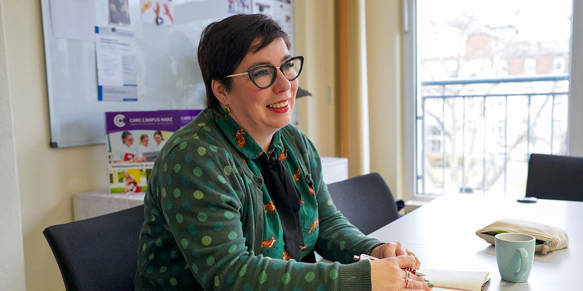 KI generiert: Eine Person mit Brille sitzt lächelnd an einem Tisch in einem Büro oder Besprechungsraum. Sie hält einen Stift und es liegt eine Tasse vor ihr.
