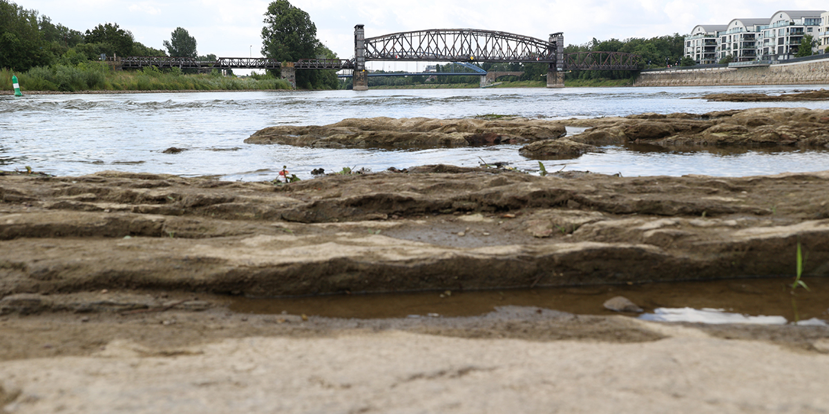 KI generiert: Das Bild zeigt eine flache, felsige Flusslandschaft mit einer Stahlbrücke im Hintergrund, die den Fluss überquert. An den Ufern sind Vegetation und Gebäude sichtbar.
