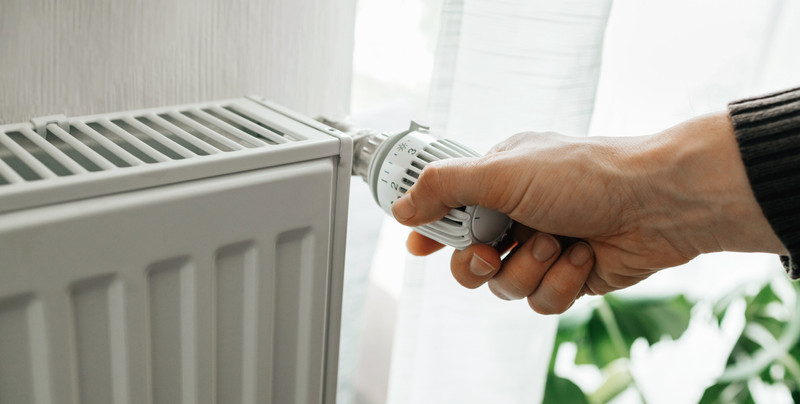 KI generiert: Eine Hand dreht den Thermostat eines Heizkörpers, um die Raumtemperatur einzustellen. Im Hintergrund ist ein Fenster mit Vorhängen zu sehen.