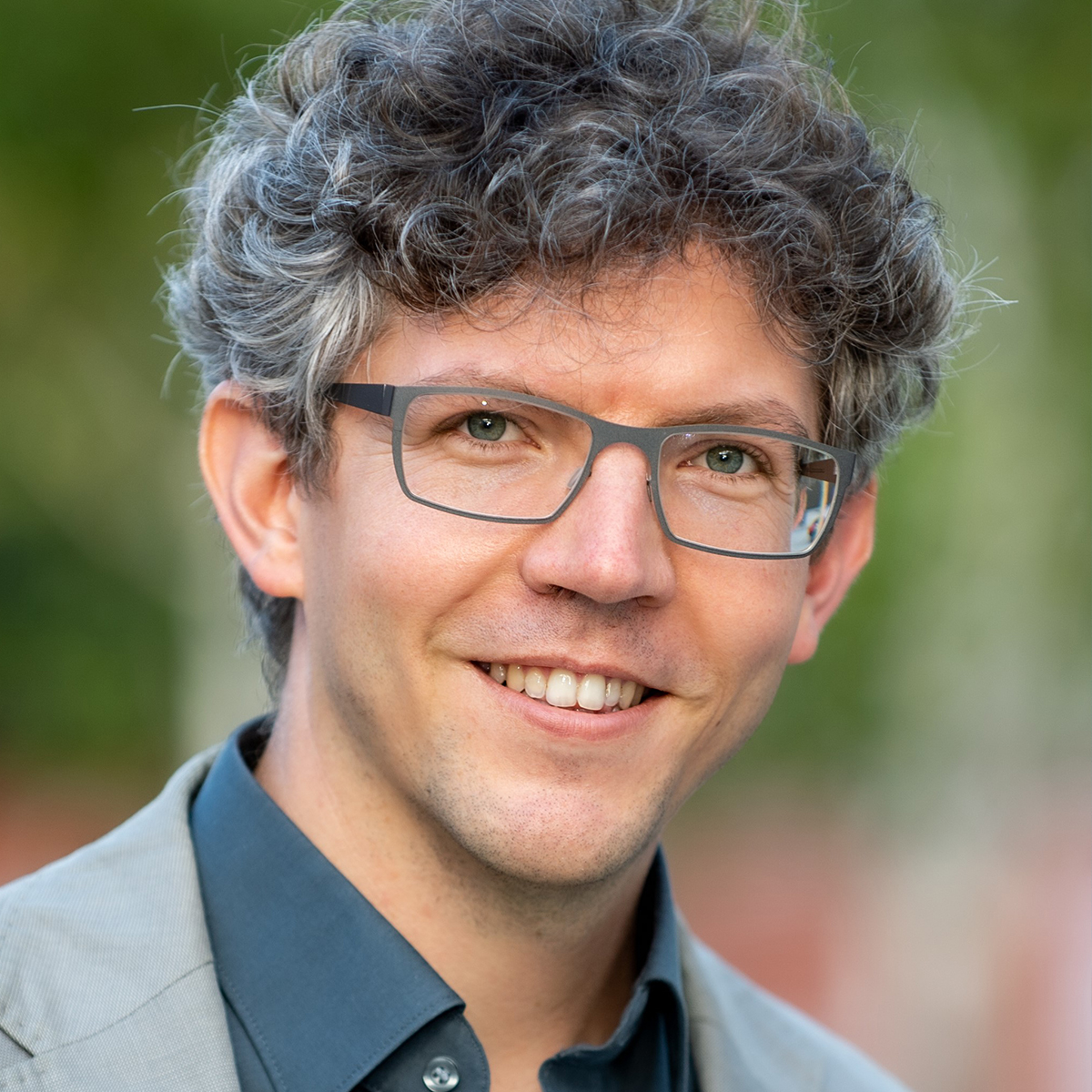 KI generiert: Das Bild zeigt einen Mann mit lockigem Haar und Brille, der lächelt. Der Hintergrund ist unscharf und grünlich.
