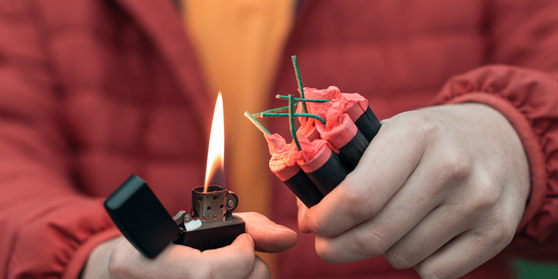 KI generiert: Das Bild zeigt eine Person, die dabei ist, eine Gruppe von zusammengesteckten Feuerwerkskörpern oder Böllern mit einem Feuerzeug zu entzünden. Die Szene fokussiert auf die Hände und den potenziell riskanten Moment des Zündens.