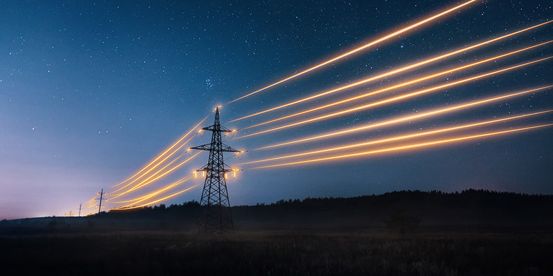 KI generiert: Das Bild zeigt Strommasten mit beleuchteten Stromleitungen, die sich diagonal durch die Landschaft erstrecken, vor einem Sternenhimmel. Die leuchtenden Linien erzeugen einen dynamischen Effekt und kontrastieren mit der nächtlichen Umgebung.