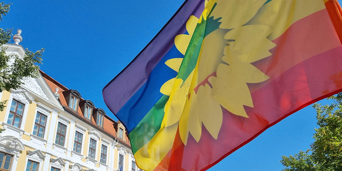 KI generiert: Das Bild zeigt eine bunte Flagge mit einem stilisierten gelben Sonnenblumenmotiv im Vordergrund, die vor einem historisch anmutenden Gebäude und einem klaren blauen Himmel weht. Der Hauptinhalt des Bildes ist die Flagge, die im Mittelpunkt steht und farbenfroh hervorsticht.