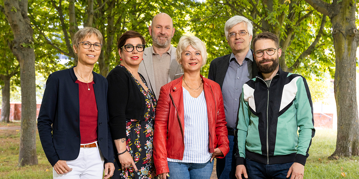 KI generiert: Eine Gruppe von sechs Personen steht lächelnd in einer von Bäumen gesäumten Allee. Die Personen tragen unterschiedliche, farbenfrohe Kleidung und posieren nah beieinander.