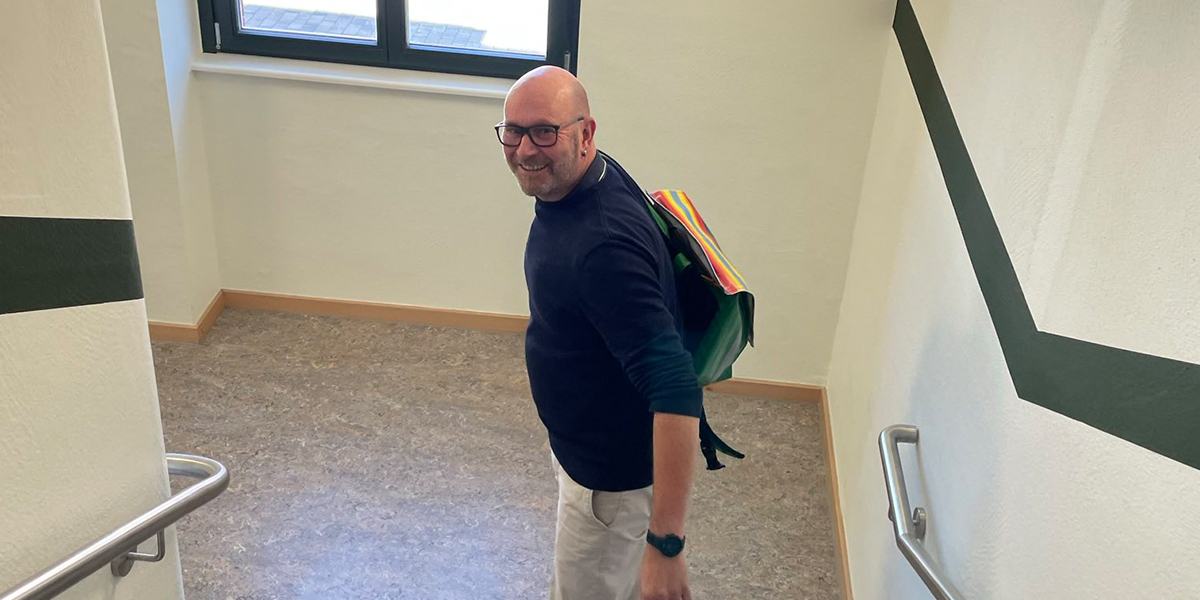 KI generiert: Ein Mann mit einem bunten Rucksack steht in einem Treppenhaus und schaut lächelnd zurück zur Kamera. Er trägt ein dunkles Oberteil und eine helle Hose.