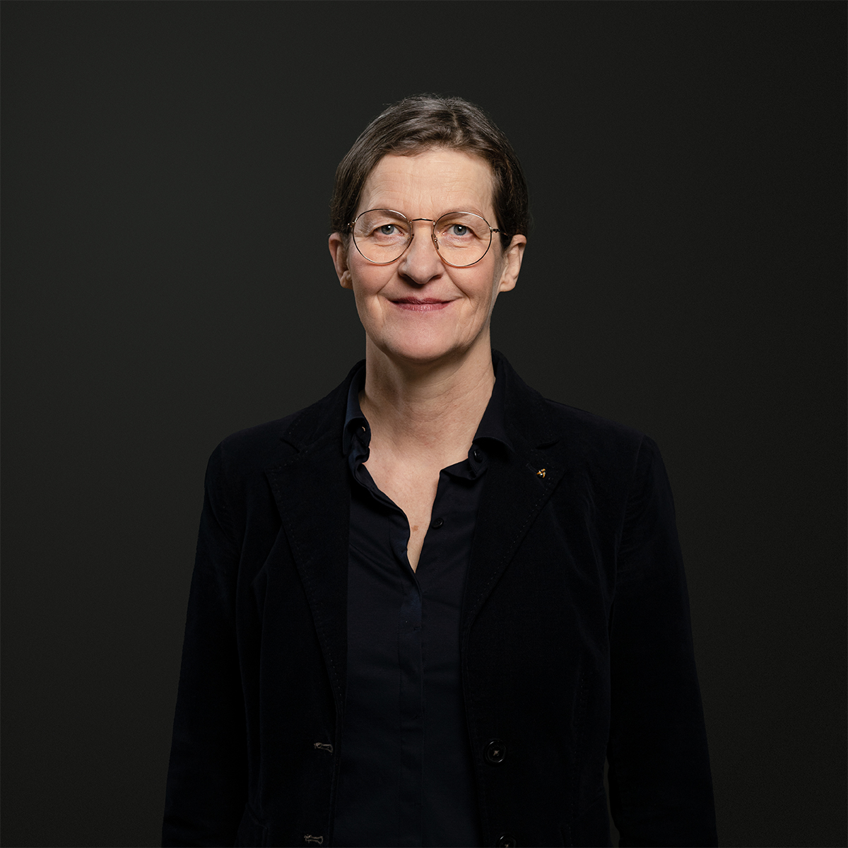 KI generiert: Das Bild zeigt eine Person, die freundlich in die Kamera lächelt. Die Person trägt eine Brille und ist in dunkler Kleidung vor einem neutralen Hintergrund abgebildet.