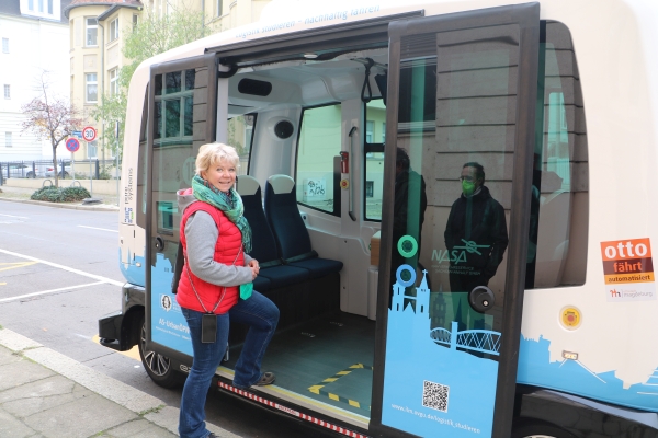 KI generiert: Eine Frau steigt in ein autonomen Kleinbus ein, der an einer Straße steht. Der Bus ist modern gestaltet und zeigt Aufkleber, die auf ein Projekt zu automatisiertem Fahren hinweisen.