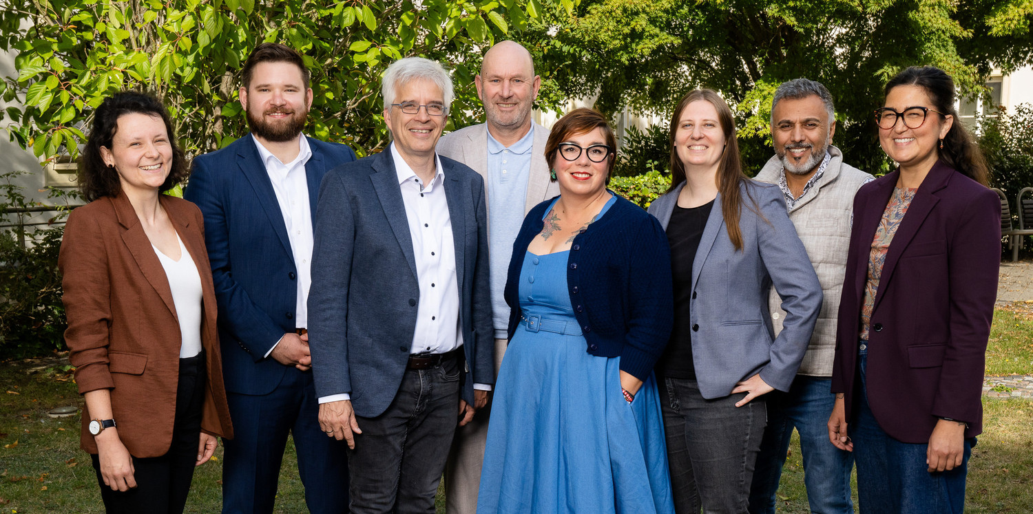KI generiert: Das Bild zeigt eine Gruppe von acht Personen, die in einem Garten oder Park stehen und sich für ein Gruppenfoto versammelt haben. Der Hauptinhalt des Bildes ist die Darstellung einer gemischten Gruppe von Menschen, die sich in einer entspannten, natürlichen Umgebung befinden.