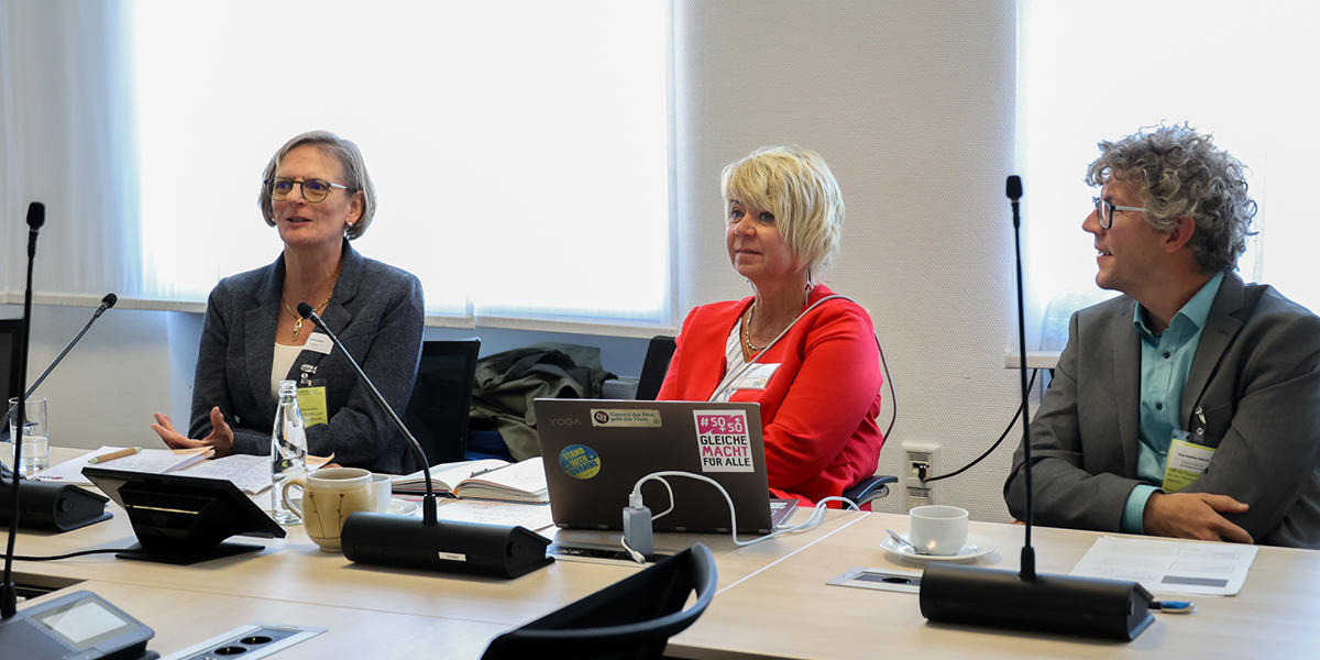 KI generiert: Das Bild zeigt drei Personen, die an einem Sitzungstisch in einem Besprechungsraum sitzen und scheinbar an einer Diskussion teilnehmen. Vor ihnen befinden sich Mikrofone, Notizen und ein Laptop mit Aufklebern.