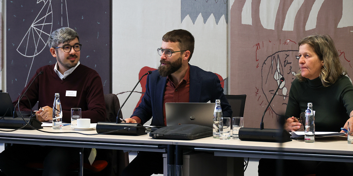 KI generiert: Drei Personen sitzen an einem Tisch mit Mikrofonen und Computern, als ob sie an einer Besprechung oder Konferenz teilnehmen. Im Hintergrund sind dekorative Paneele zu sehen.