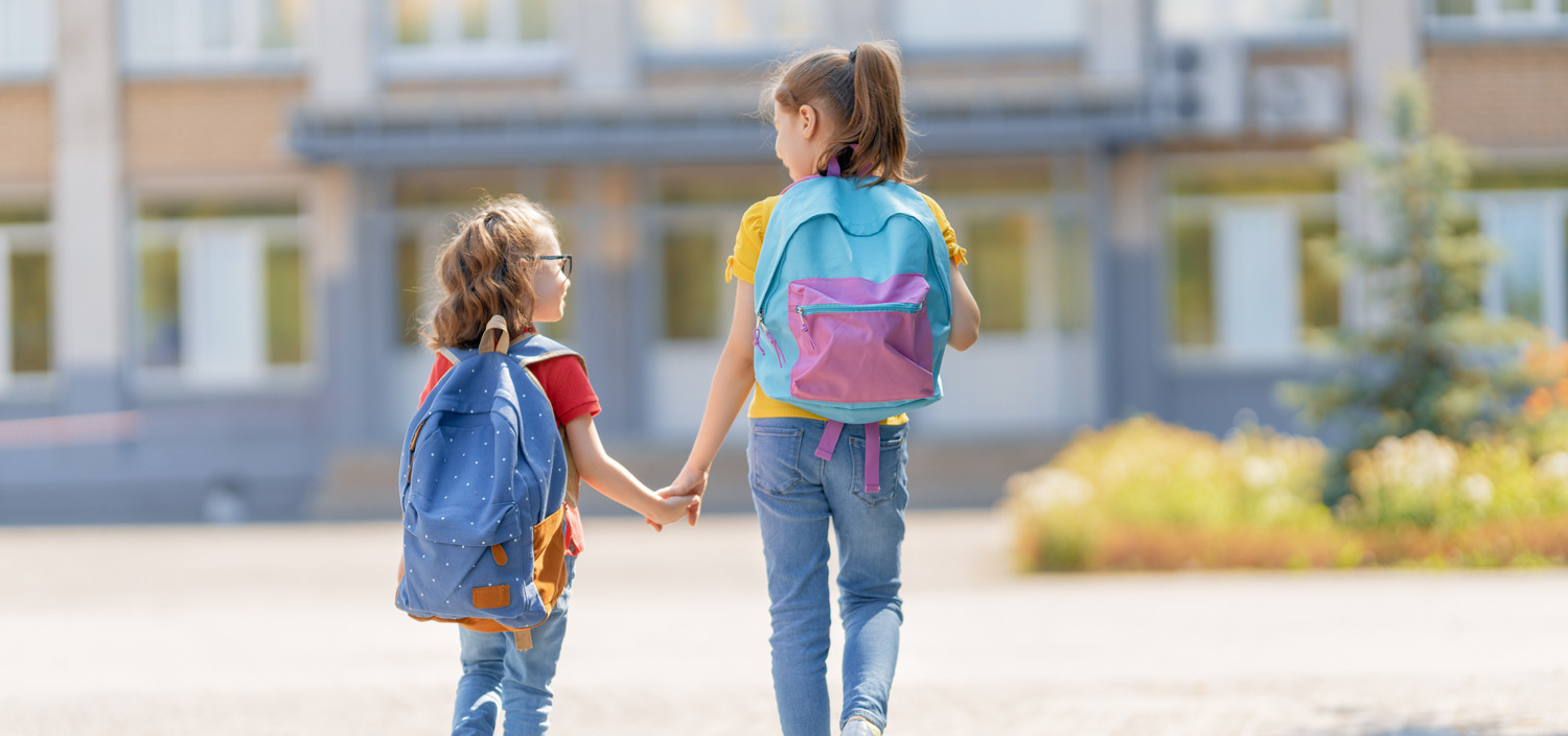 KI generiert: Das Bild zeigt zwei Kinder mit Rucksäcken, die sich an der Hand halten und auf ein Schulgebäude zugehen. Sie scheinen auf dem Weg zur Schule zu sein.