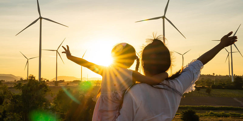 KI generiert: Das Bild zeigt einen Sonnenuntergang hinter mehreren Windturbinen, während eine erwachsene Person und ein Kind mit erhobenen Armen die Aussicht genießen. Der Hauptinhalt des Bildes ist die Verbindung von Mensch und erneuerbarer Energie in einer natürlichen Umgebung.