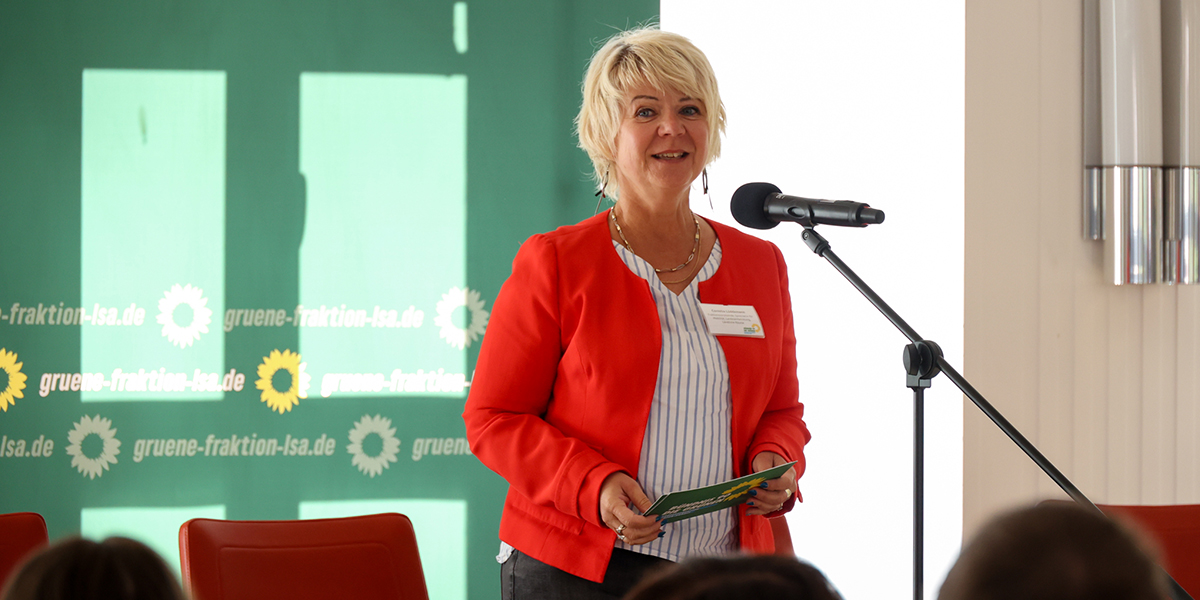 KI generiert: Das Bild zeigt eine Frau in einem roten Blazer, die vor einem Mikrofon spricht und lächelt. Im Hintergrund ist ein grünes Banner mit dem Logo und der Website der Grünen Partei zu sehen.