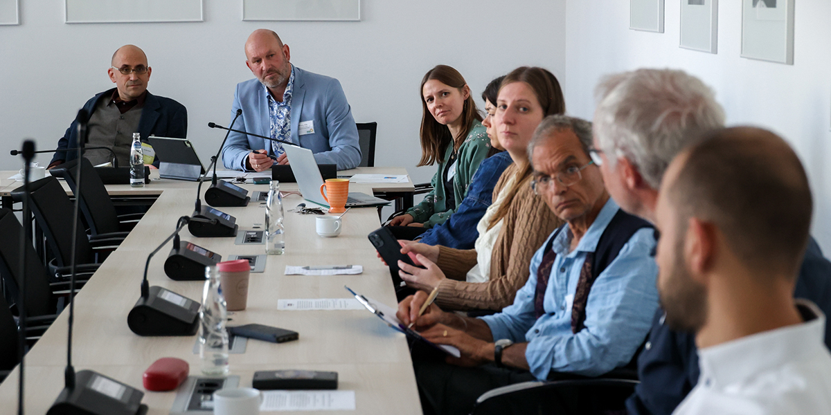 KI generiert: Das Bild zeigt eine Gruppe von Personen, die an einem Tisch in einem Besprechungsraum sitzen und ein Gespräch führen. Im Mittelpunkt stehen Diskussion und Zusammenarbeit.