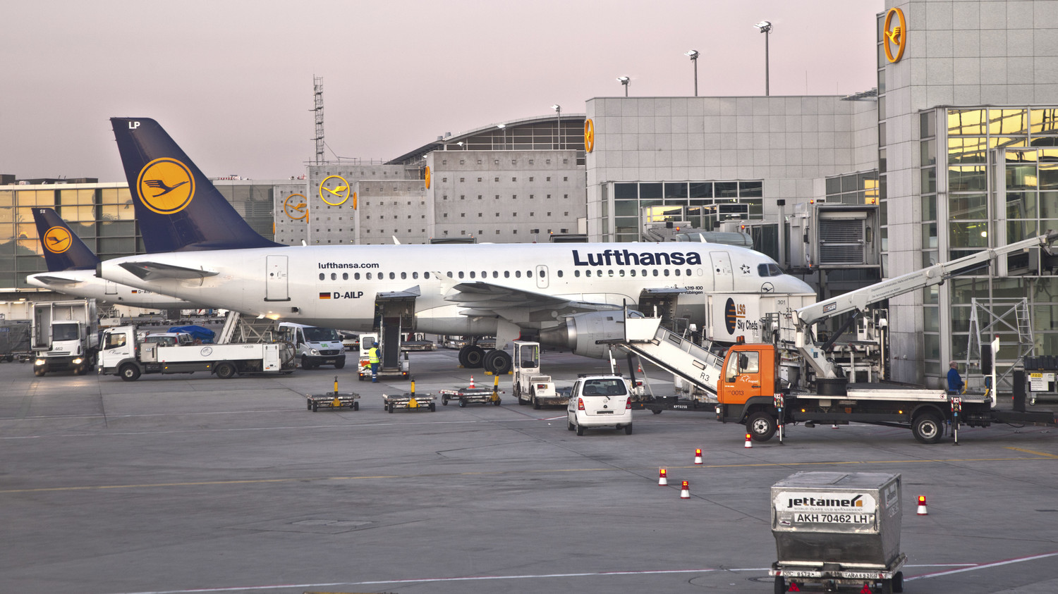 KI generiert: Das Bild zeigt ein Lufthansa-Flugzeug, das an einem Gate eines Flughafens steht. Im Vordergrund sind verschiedene Bodenfahrzeuge und Gepäckwagen zu sehen, die am Flugzeug arbeiten.