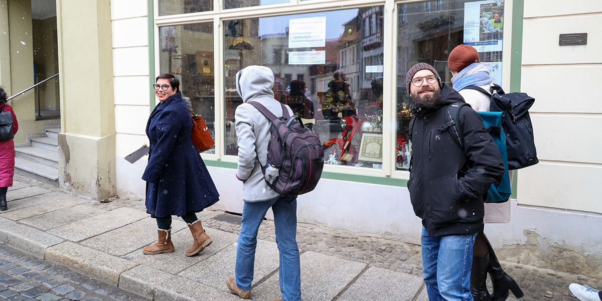 KI generiert: Das Bild zeigt eine Gruppe von Menschen, die auf einem Gehweg vor einem Geschäft mit verzierten Fenstern stehen. Es ist winterlich, einige tragen warme Kleidung und es scheint leicht zu schneien.