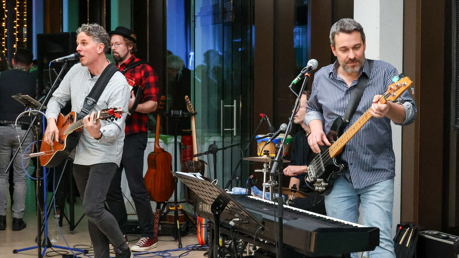 KI generiert: Eine Band tritt in einem Innenraum auf, wobei der Fokus auf drei Musikern liegt, die Gitarre und Bass spielen und singen. Im Hintergrund sind Instrumente und weitere Bandmitglieder zu sehen.