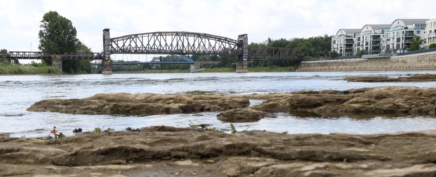 KI generiert: Das Bild zeigt eine Flusslandschaft mit einer markanten Eisenbahnbrücke im Hintergrund. Im Vordergrund sind flache Felsen im Wasser zu sehen.