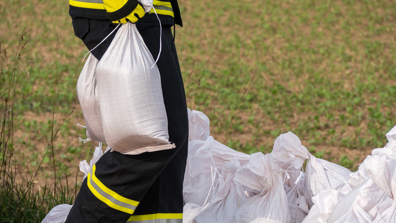 KI generiert: Das Bild zeigt eine Person in einer Feuerwehruniform, die Sandsäcke trägt. Im Hintergrund sind weitere aufgestapelte Sandsäcke vor einem Feld zu sehen.