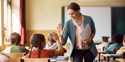 KI generiert: Das Bild zeigt eine Lehrerin in einem Klassenzimmer, die mit einem Schüler einen High-Five gibt. Im Hintergrund sind weitere Schüler zu sehen, die an ihren Schreibtischen sitzen.