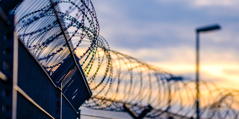 KI generiert: Das Bild zeigt eine Nahaufnahme von Stacheldraht auf einem Zaun bei Sonnenuntergang. Der Fokus liegt auf den scharfen Kanten des Drahtes, während der Hintergrund sanft verschwimmt.