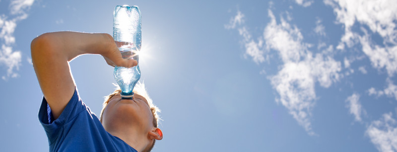 KI generiert: Ein Junge trinkt an einem sonnigen Tag Wasser aus einer Plastikflasche. Der Himmel ist klar mit einigen Wolken.