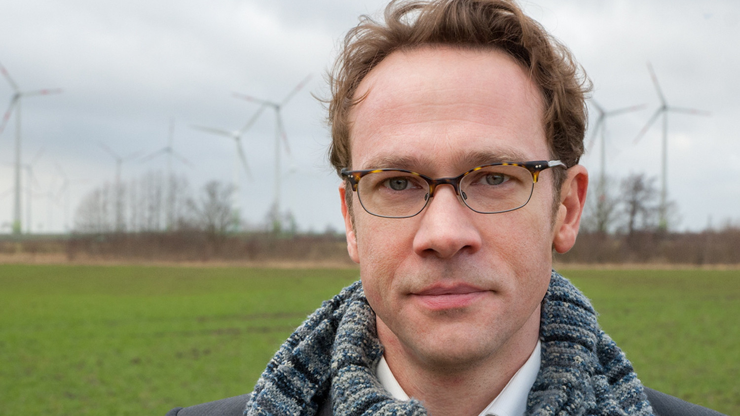 KI generiert: Ein Mann mit Brille steht im Vordergrund und trägt einen Schal, während im Hintergrund mehrere Windkraftanlagen auf einem Feld zu sehen sind. Das Bild scheint eine Verbindung zwischen Mensch und erneuerbarer Energie herzustellen.