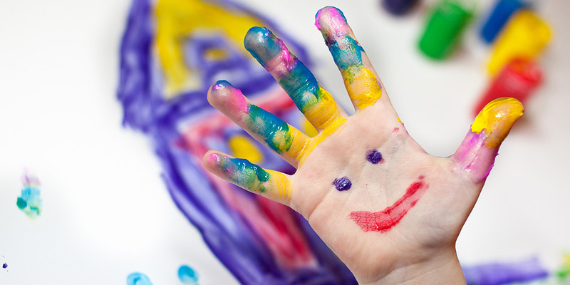 KI generiert: Das Bild zeigt eine Hand, die mit bunten Fingerfarben bemalt ist, wobei ein lächelndes Gesicht auf die Handfläche gemalt wurde. Im Hintergrund sind verschwommene Farbkleckse und ein gemaltes Bild zu sehen.