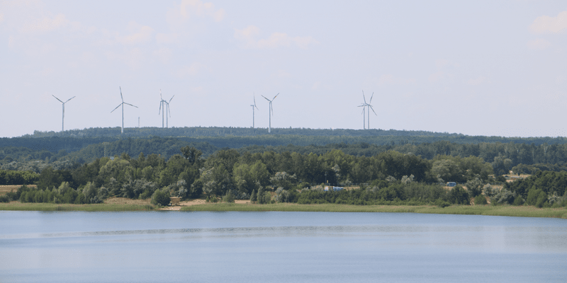 KI generiert: Das Bild zeigt eine Landschaft mit einem Gewässer im Vordergrund und einem Wald im Hintergrund, in dem mehrere Windkraftanlagen zu sehen sind. Der Hauptinhalt des Bildes sind die Windkraftanlagen auf einem bewaldeten Hügel.