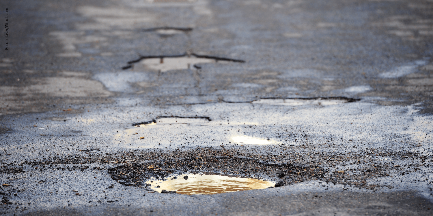 KI generiert: Das Bild zeigt eine Straße mit mehreren Schlaglöchern, in denen sich Wasser gesammelt hat. Die Oberfläche ist sichtlich uneben und beschädigt.