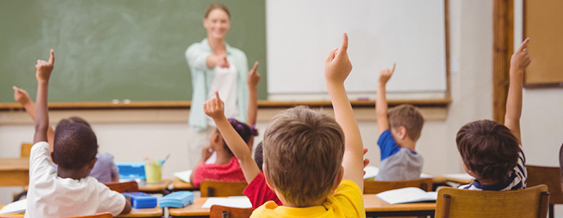 KI generiert: Das Bild zeigt eine Lehrerin, die vor einer Gruppe von Schülern in einem Klassenzimmer steht. Die Schüler heben die Hand, wahrscheinlich um eine Frage zu beantworten oder um Aufmerksamkeit zu erhalten.