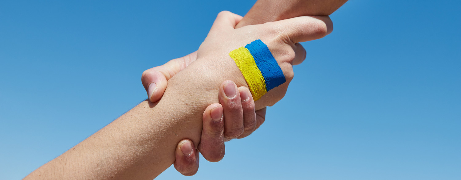 KI generiert: Das Bild zeigt zwei Hände, die sich festhalten, wobei eine Hand ein blau-gelbes Armband trägt. Der Hauptinhalt des Bildes ist die symbolische Darstellung von Unterstützung oder Solidarität, möglicherweise in Bezug auf die Ukraine, deren Nationalfarben Blau und Gelb sind.